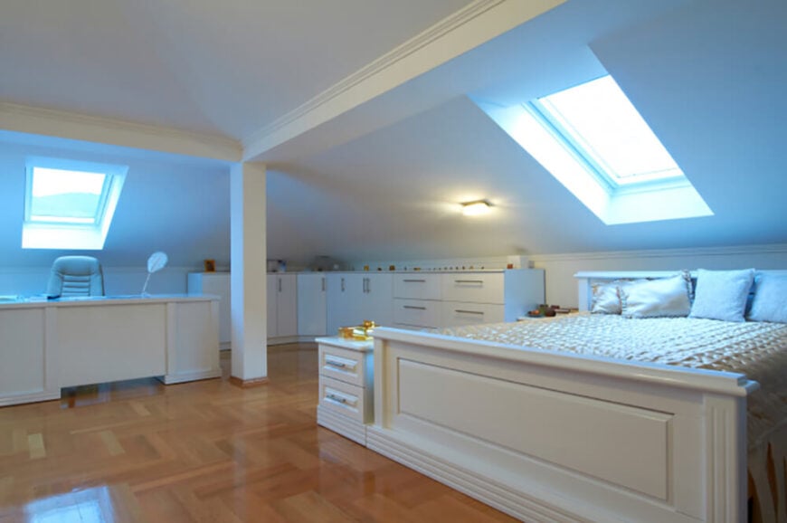 这间卧室整洁而现代，有干净的白色墙壁和家具。倾斜的天花板表明这间卧室位于房子的顶部附近，天窗让自然光涌入，照亮了床和书桌区域。硬木地板上有独特的图案，具有高光泽，有助于反光。