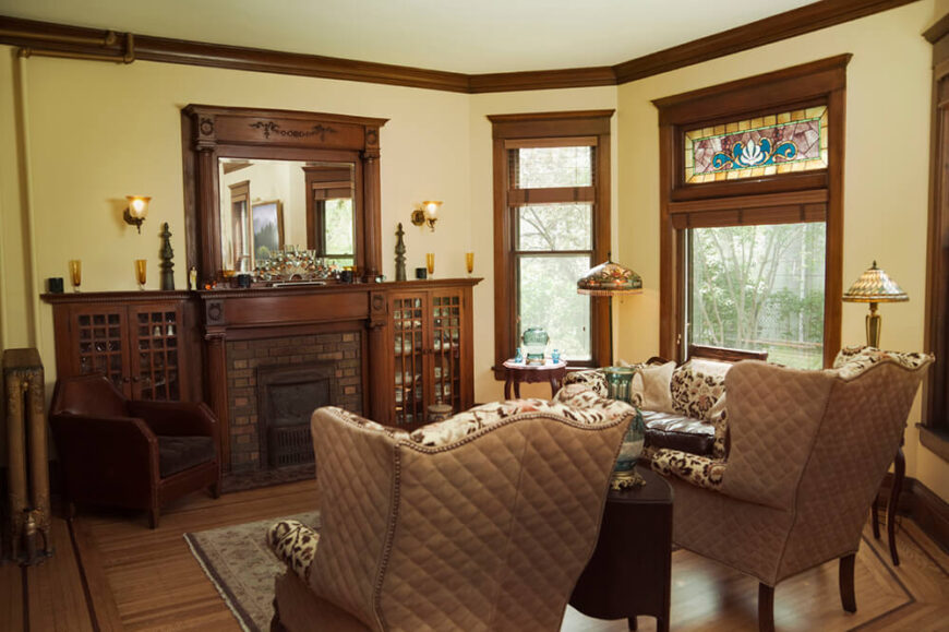 这间客厅设有大型软垫椅子，以及令人印象深刻的壁炉和壁炉。窗户周围的深色木材装饰有助于为空间设定总体基调。最大的窗户上方有一小部分彩色玻璃，为这个房间增添了色彩。