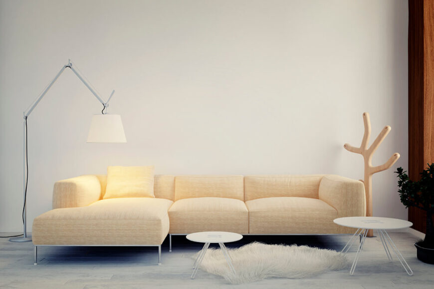一个独特和极简主义的设计，围绕着一个轻卡其色沙发和羽毛毛绒地毯。两个白色的边桌为空间增添了功能，落地灯和木制帽架衬托着沙发。
