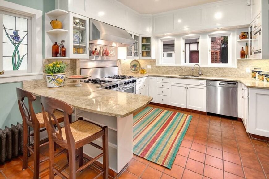 这间厨房的地板上铺着生锈的棕色瓷砖，还有白色的橱柜和米黄色的纹理台面。在餐厅扩建部分的旁边，有一个花卉图案的彩色玻璃窗。