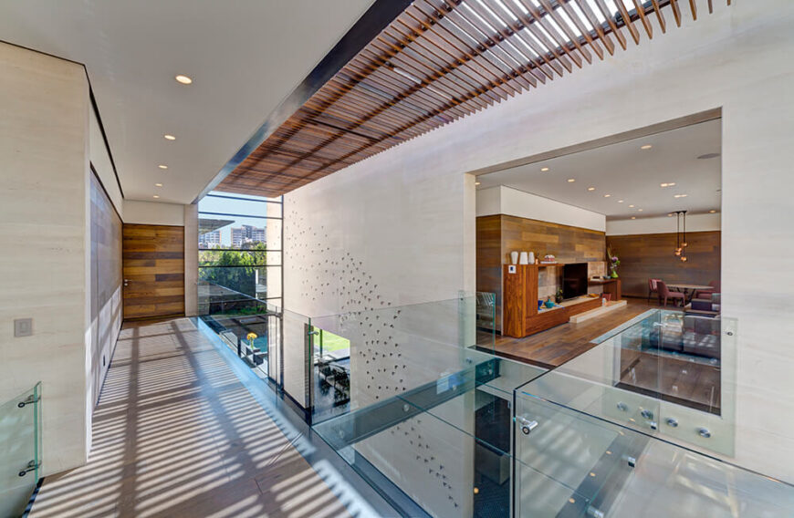 上方的木质百叶面板将阳光漫射到整个开放的大厅空间，两端的通高玻璃取代了它。在右边，一个舒适的客厅空间被丰富的木镶板所定义。