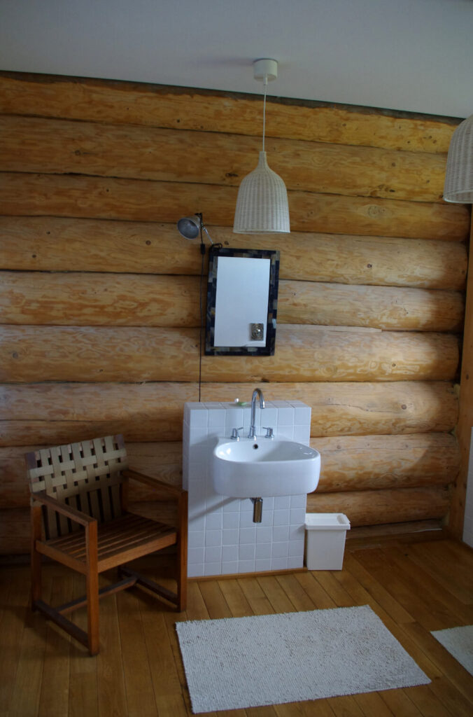 这是其中一间浴室，又是一种优雅的克制。浮动水槽安装在原木墙边的一小块白色瓷砖上，上面只有一面小镜子和一对吊灯，旁边还有一把木椅。