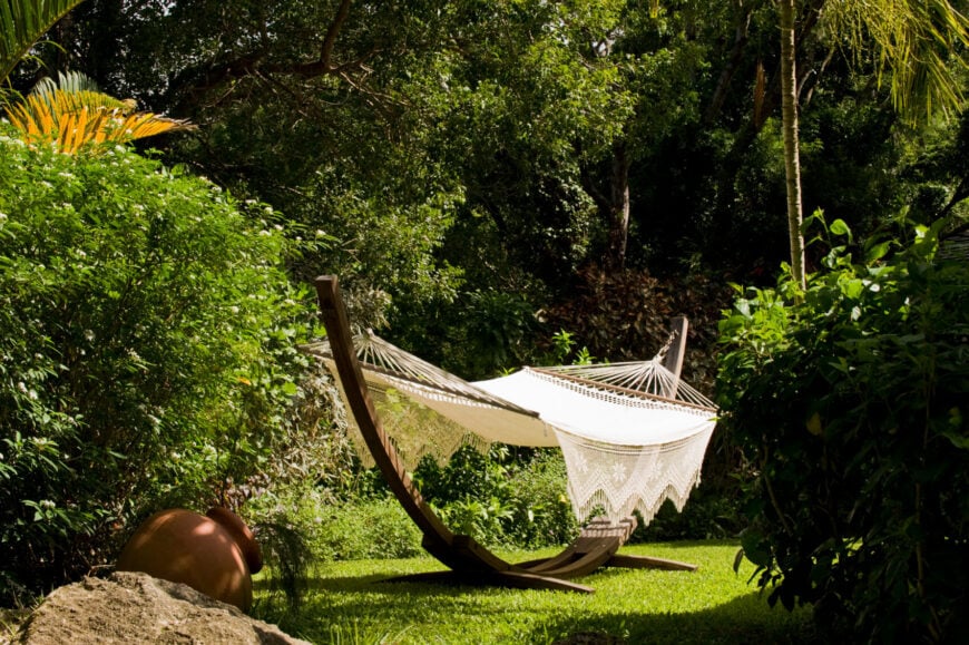 这张可爱的钩针吊床挂在一个半圆木制支架的两端。吊床坐落在院子里树木丛生的僻静处。