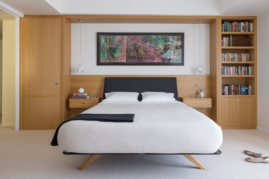卧室延续了丰富的木材色调和现代极简主义风格的融合，一个墙壁大小的架子和存储单元支撑着光滑而现代的床。墙上挂着一幅彩色的艺术品，代替了床头板。