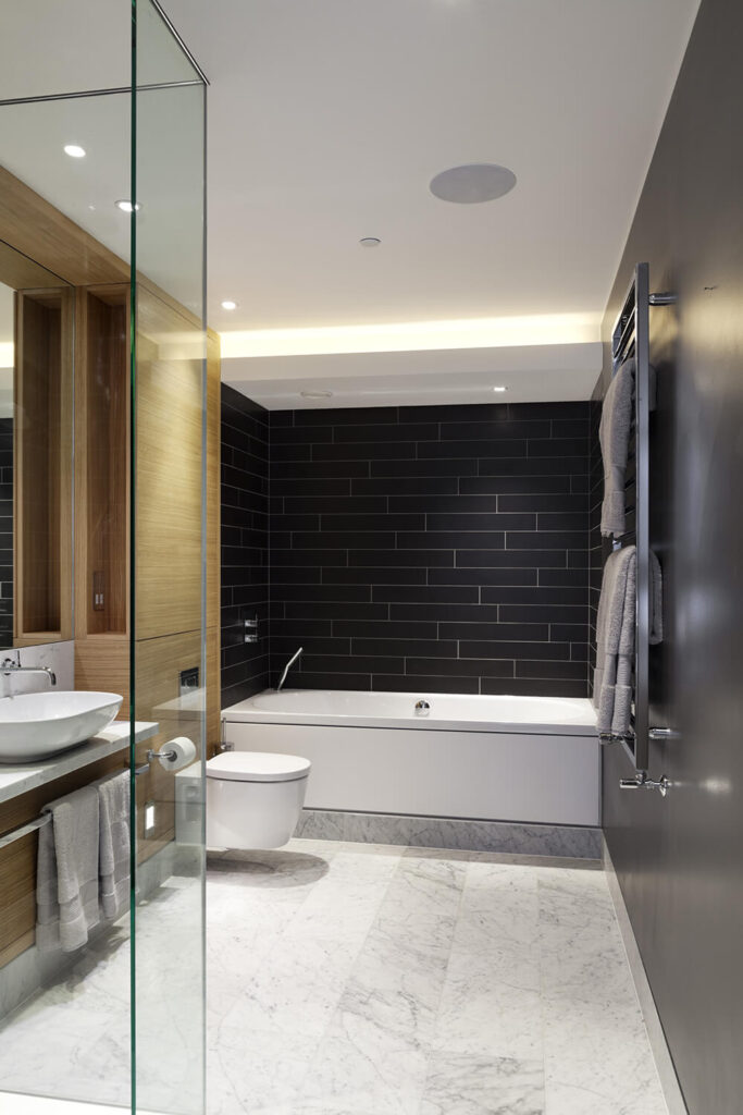 浴室充满了强烈的对比，有触感的元素，有黑色的瓷砖墙壁，丰富的天然木橱柜，大理石地板，和一个带水槽的浮动梳妆台。