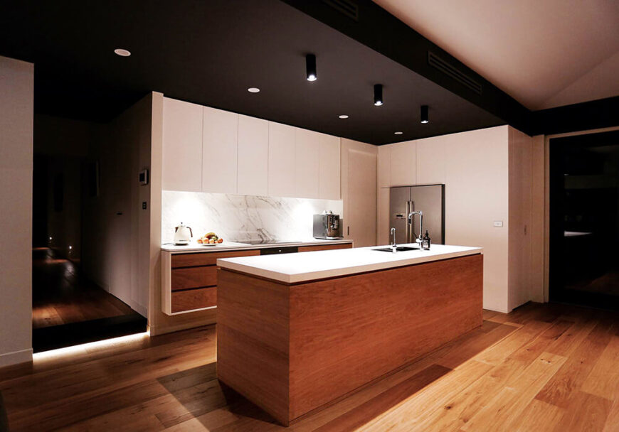 现在让我们仔细看看厨房，这是一个光滑的极简主义空间，由一个带有白色台面的大岛台主导。微妙的白色大理石后挡板增加了一剂优雅，而光滑的白色橱柜强调了设计的克制。