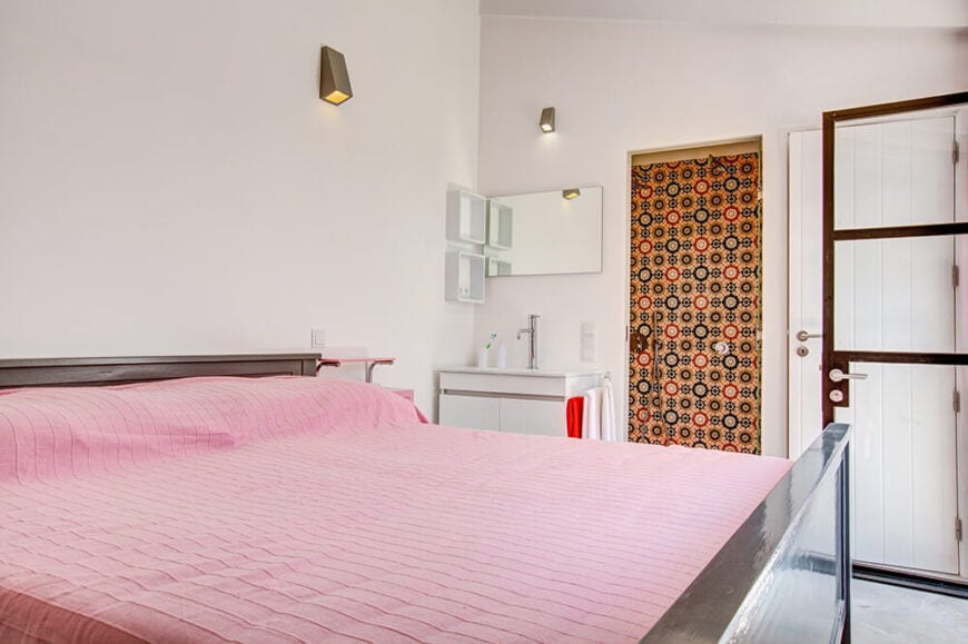 这个副卧室以一个巨大的现代床架为中心，床架上有淡淡的粉红色床单。连接的浴室通过一扇高度图案的黄色门进入，旁边是一个白色的小梳妆台。