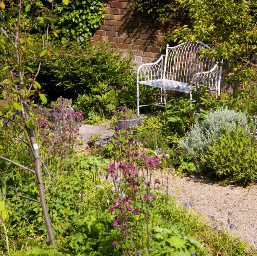 这是一个简单的金属长凳，被漆成白色。它没有与周围环境融为一体，而是给该地区带来了一种轻盈。这个花园与绿色植物形成了鲜明的对比。