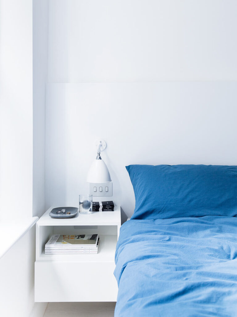 主卧室唯一的色彩是大胆的蓝色床罩，旁边的墙上有一个漂浮的白色床头柜。大窗户能提供最大程度的被动照明。
