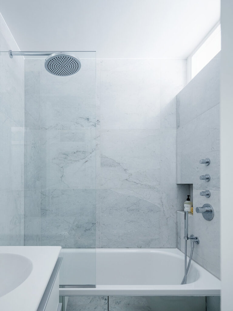 主浴室采用白色大理石，为白色极简主义外观增添了浓郁的质感元素。一个大的浴缸是用玻璃框起来的，而上面的一扇纤细的窗户为房间提供了一些阳光。