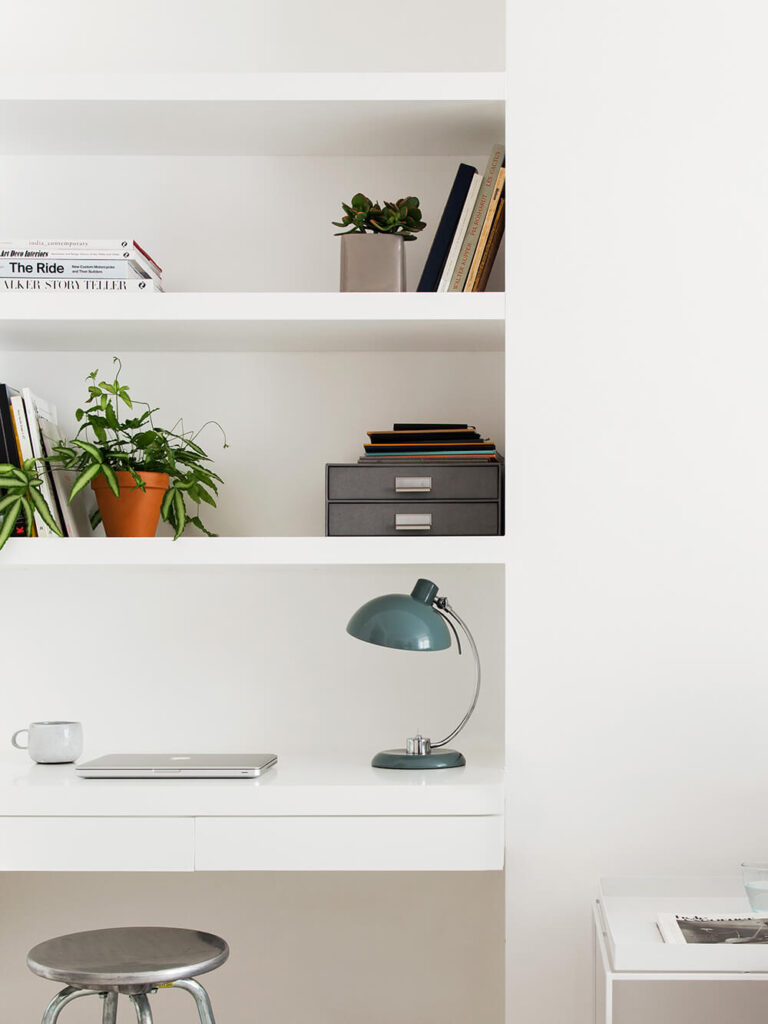 舒适的家庭办公空间设有内置办公桌和丰富的书架，全部为白色，与墙壁完美融合。色彩和镀铬的微妙实例突出了极简主义的调色板。