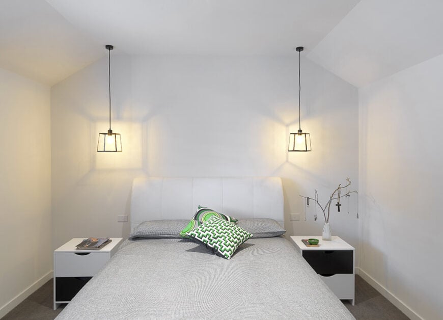 主卧室与开放式客厅和餐厅区采用了相同的简约风格。白色的墙壁，灰色的地毯和灰色色调的床，两侧是一对白色的床头柜和金属框架吊灯。
