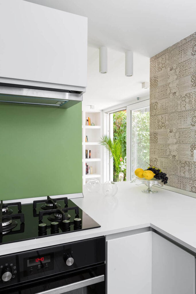 走到柜台的另一边，我们看到厨房被光滑的白色表面包裹着，极简主义的台面和橱柜框架光滑的黑色和不锈钢电器。大胆的绿色色调和纹理花卉墙纸增强了外观。