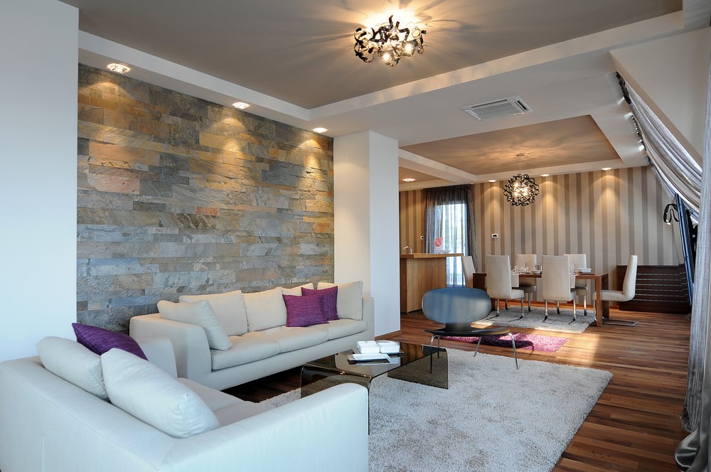 木地板和大理石墙壁在传统的窗帘和暖色调的氛围中增添了现代的感觉。由玻璃制成的简单而朴素的中央桌子，配有紫色软垫枕头的素色布艺沙发，都是这个设计中的当代风格。