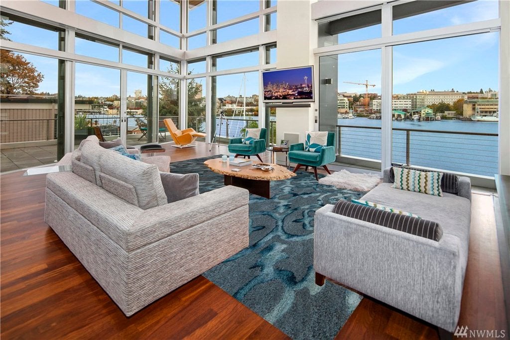 一个布置精美的客厅，可以看到西雅图的蓝色海水。家具图案统一，与透明玻璃墙外的蓝色主题环境相呼应。它更像是一个现代的室内装饰，有几个色彩鲜艳的座椅和枕头。