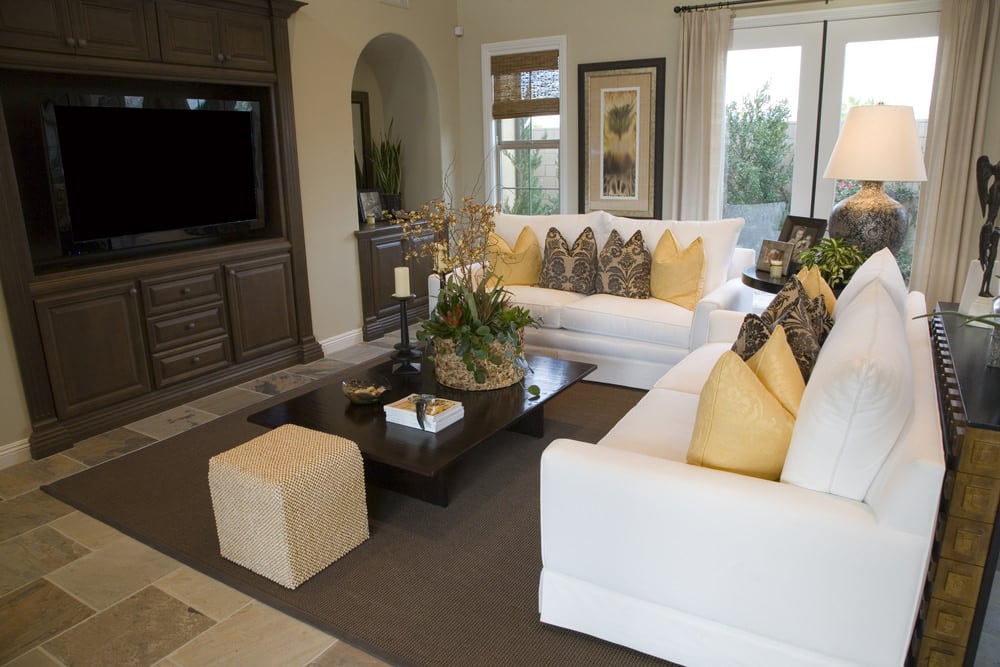纯白沙发搭配金色和黑色图案的装饰性抱枕，突显了这个老式的、受自然启发的客厅。咖啡桌的两侧放着一个立方体软脚凳。