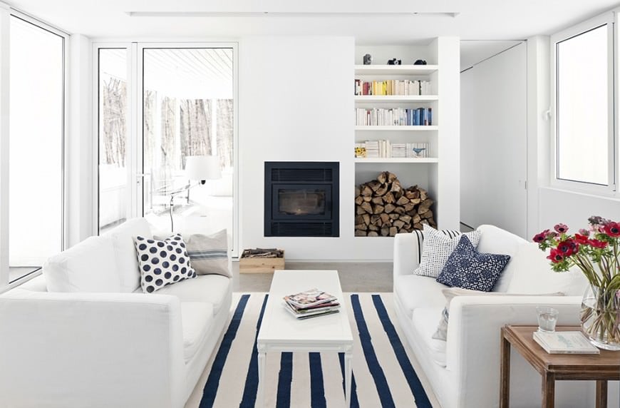 客厅布置得很好，白色家具配以蓝色条纹地毯和花纹抱枕。白色的架子上堆着柴火，角落里的木桌子上点缀着一些鲜花，让人在纯白的风景中转移了一点注意力。