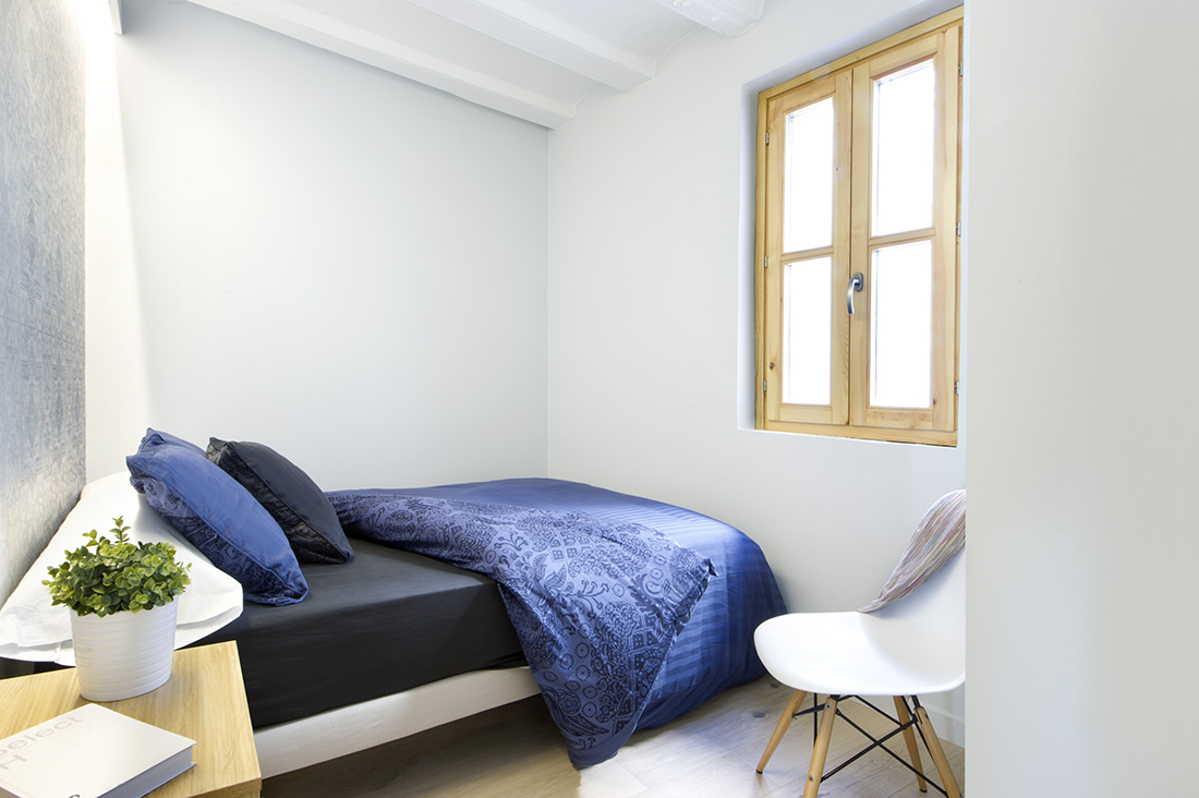 一个小而自由移动的卧室，有一个柔软的床垫，暗蓝色的被子和枕头。它还有一个普通大小的窗户，用于通风和自然采光。