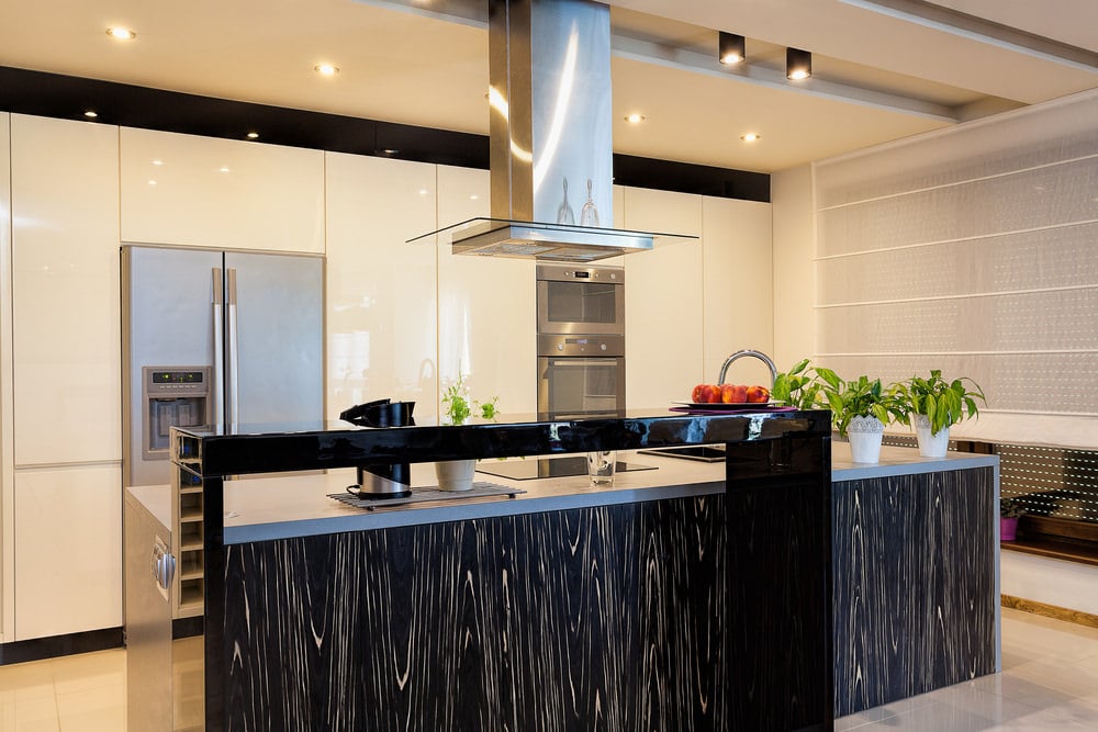 时尚的厨房提供了一个不锈钢通风罩，悬挂在一个时尚的厨房岛框上，边框上有凸起的高光泽黑色柜台。
