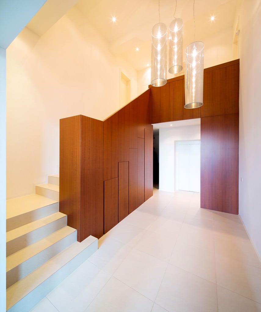 极简主义风格的入口，白色瓷砖地板和楼梯，配有实木栏杆，配有内置储物柜。它由花哨的圆柱形吊坠和安装在白色天花板上的凹形灯照亮。