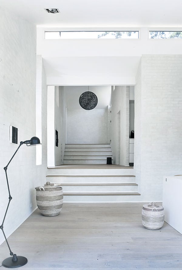 斯堪的那维亚风格的门厅，白色砖墙和混凝土楼梯配有木踏板。它由一盏黑色落地灯和一个编织的球形吊坠照亮。