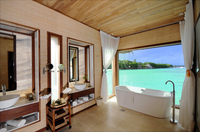 热带主浴室设有一个独立的浴缸，前面有一个大窗户，可以看到海洋的景色。这个空间还包括木质天花板、瓷砖地板和带水槽的梳妆台。