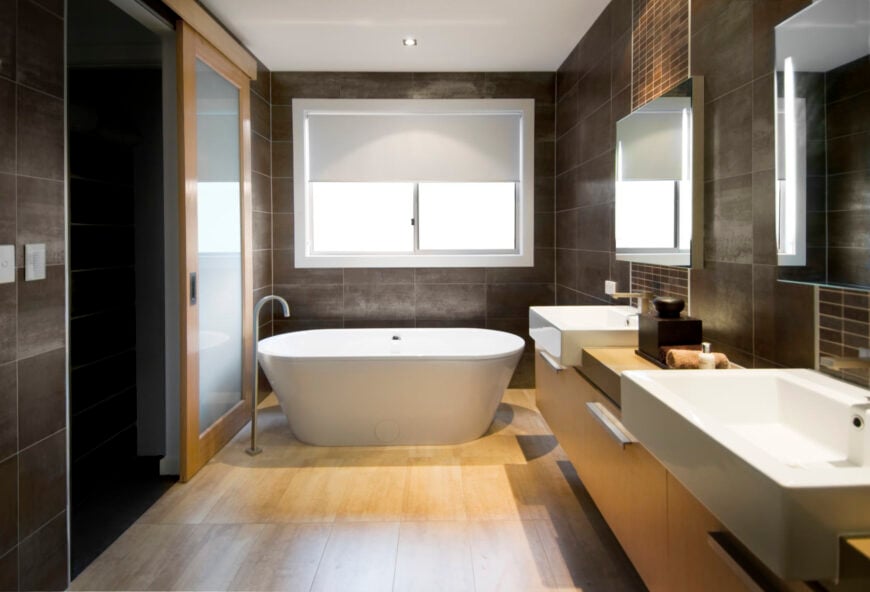 现代化的主浴室设有一个白色的独立浴缸，周围环绕着棕色瓷砖墙和硬木地板。一面墙上有一个漂浮的木制梳妆台，上面有两个水槽。