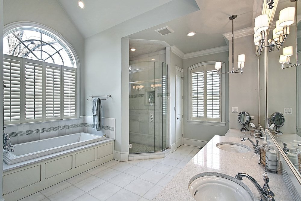 白色浴室，铺着瓷砖地板，拱形玻璃窗上装有百叶窗。它由悬挂的灯和安装在镜子上的壁灯照明。