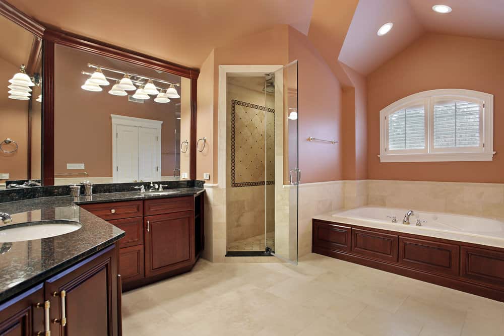 大的主浴室，橙色的墙壁和米黄色的瓷砖地板。在浴室的角落里有一个角落淋浴间。旁边是浴缸。水槽柜台拥有花岗岩台面。