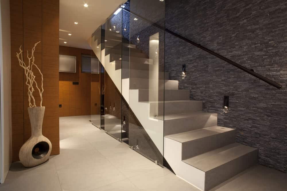 优雅的门厅展示了优雅的木制装饰和白色楼梯，从地板到天花板的玻璃板作为它的栏杆。它由安装在灰色石墙上的烛台照明。