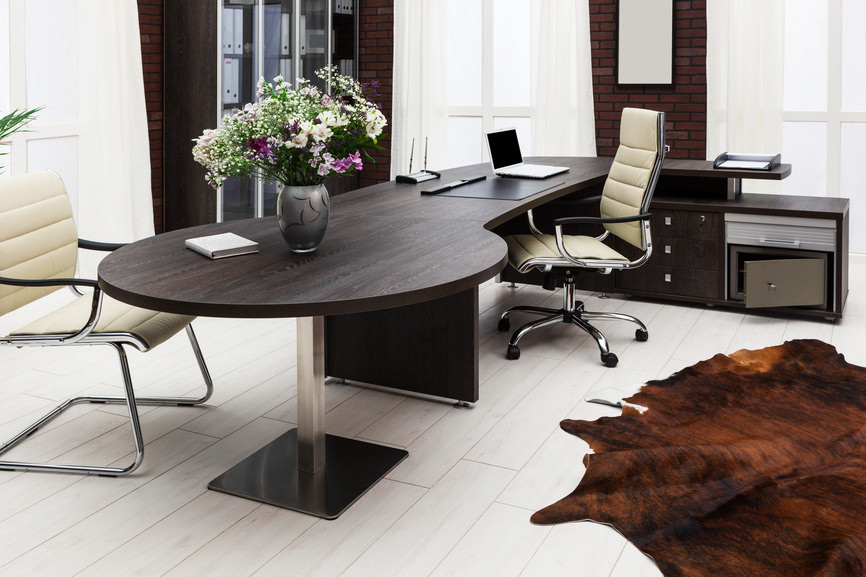 现代家庭办公室，深色木材l型办公桌，配有圆桌，顶部有一个花瓶。它有砖墙和铺在木板地板上的牛皮地毯。