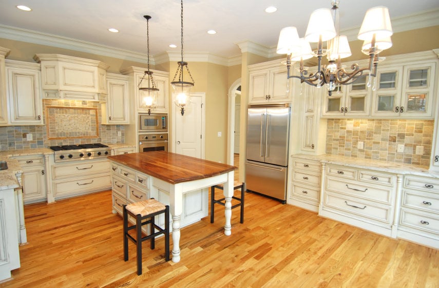 米色厨房配有吊灯、凹形和吊灯、白色橱柜、石瓷砖后挡板，以及白色底座橱柜厨房岛台，木质表面位于中色调木地板上。