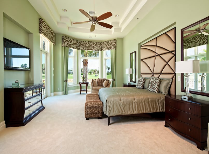 这间宽敞的主卧室设有一张舒适的床，配有镶在墙上的时尚定制床头板。它有地毯地板和一个飘窗，上面装饰着斑马印花的窗帘和与墙壁相配的绿色窗帘。