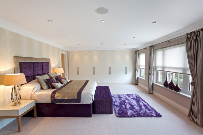 在这间米色的主卧室里，一张紫色的床和旁边的软垫长椅和地毯格外显眼。它包括一个通高的衣柜和木制床头柜，台灯照亮了房间，自然光通过玻璃窗照射进来。