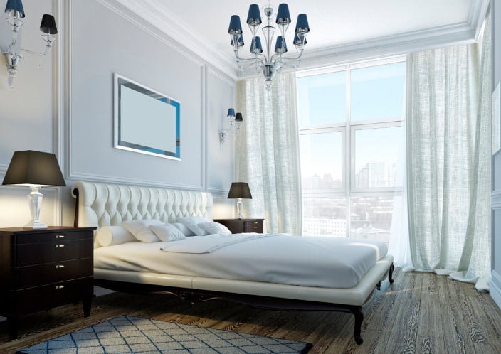自然光线透过主卧室的通高玻璃窗，展示了蓝色吊灯和配套的烛台，木质地板上的白色簇绒床。