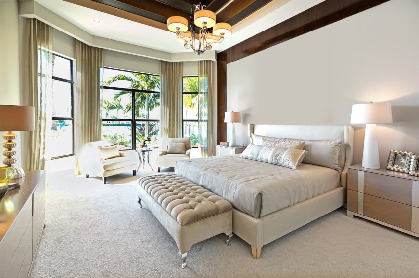 一间现代的主卧室，配有豪华的托盘天花板，由一盏令人惊叹的时尚吊灯照亮。房间里有一张大床，靠近窗户的休息区和地毯地板。