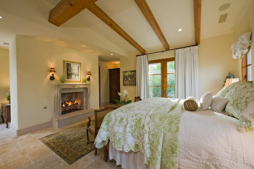 质朴的主卧室，石头瓷砖地板上有混凝土壁炉，上面有绿色地毯。它还有木制框架的窗户和白色窗帘覆盖的门。