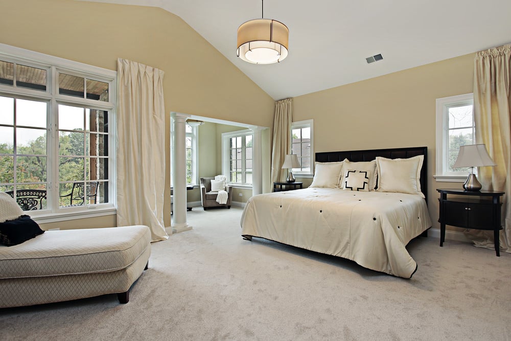 大型主卧室设有白色地毯地板和一张大床，周围是米黄色的墙壁，由华丽的天花板照明。