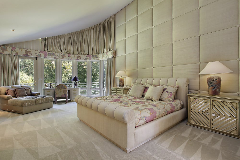 精致的主卧室拥有米黄色的簇绒墙和有图案的地毯地板。它包括一张相同色调的软垫床，上面装饰着与窗帘头部相匹配的花卉床品。