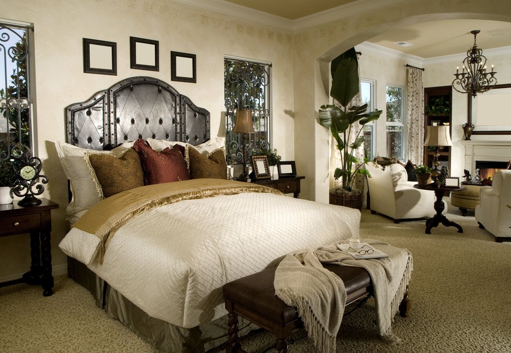 这间主卧室展示了一张皮革床，床的一端有一张长凳，休息区提供白色扶手椅，落地灯和老式枝形吊灯照明。