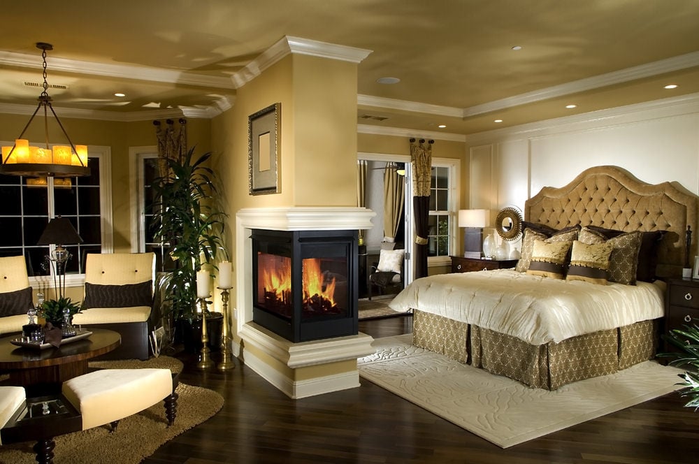 温暖的主卧室以玻璃包围的壁炉为特色，壁炉安装在米黄色的墙壁上，墙壁上有白色的冠状装饰，作为生活空间的分隔物。