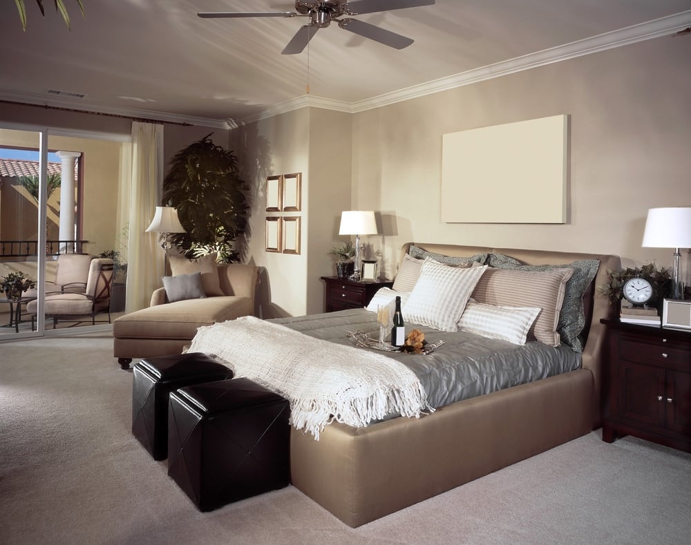 简单的主卧室里有一张米黄色的床，上面装饰着灰色的床品，与旁边的躺椅相匹配。它用一张空白的画布装饰，安装在床的上方，另一面墙上有画廊的框架。