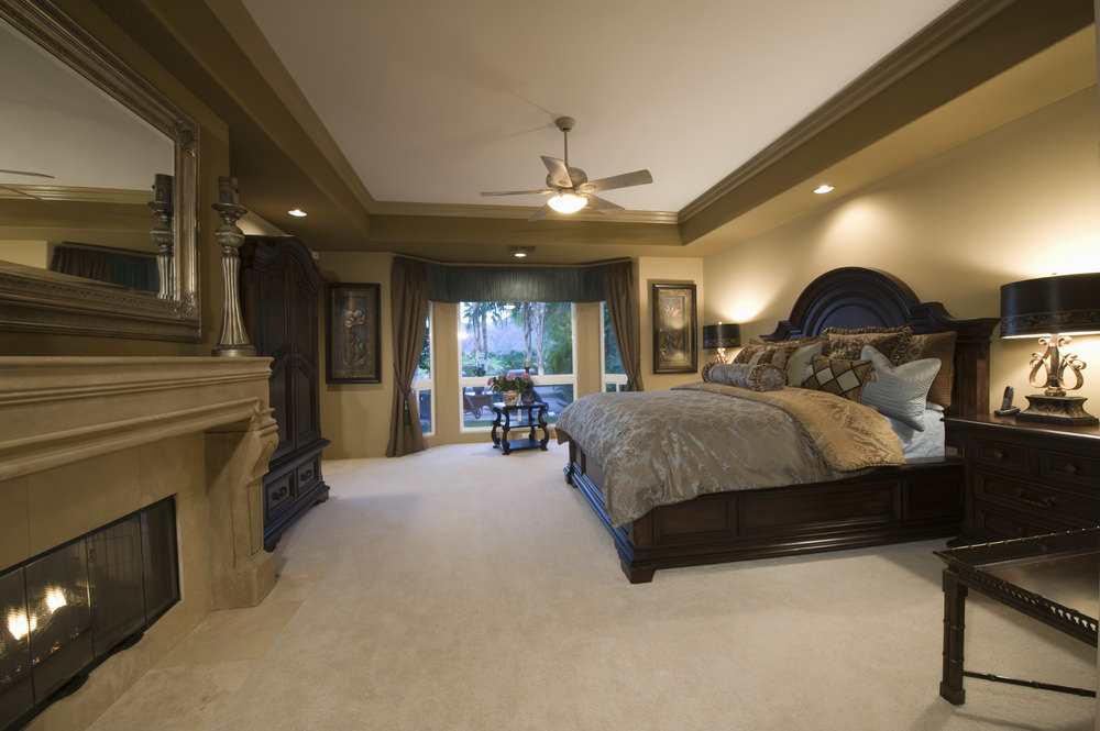 主卧室以棕色、米色和白色的色调装饰，设有一张特大号床，一个燃木壁炉和地毯地板。房间的深色木材和较浅的颜色形成对比，浅棕色的边缘环绕着白色的托盘天花板。