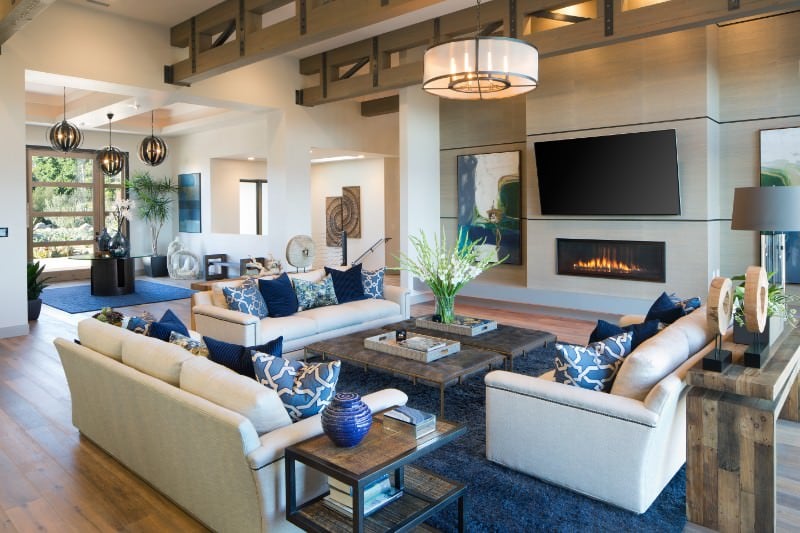现代客厅设有白色沙发，蓝色地毯覆盖硬木地板。宽敞的空间还包括大量的艺术收藏品、燃气壁炉上方的宽屏电视和带有外露横梁的天花板。