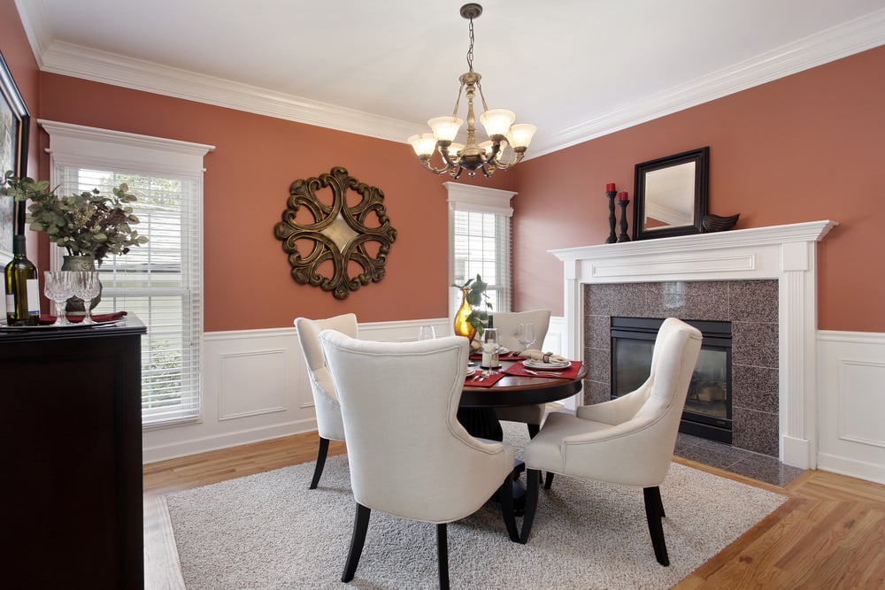 迷人的餐厅装饰着一个圆形的木头墙壁艺术安装在珊瑚粉色的墙。它有一个圆形的餐桌周围用白色后卫椅子坐在灰色的地毯,在壁炉的旁边。