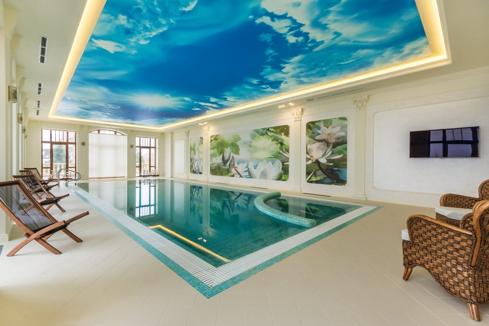 室内泳池上方的托盘天花板上有一个充满云的蓝天的图像。该空间还包括木制和藤椅，以及壁挂式平板电视。