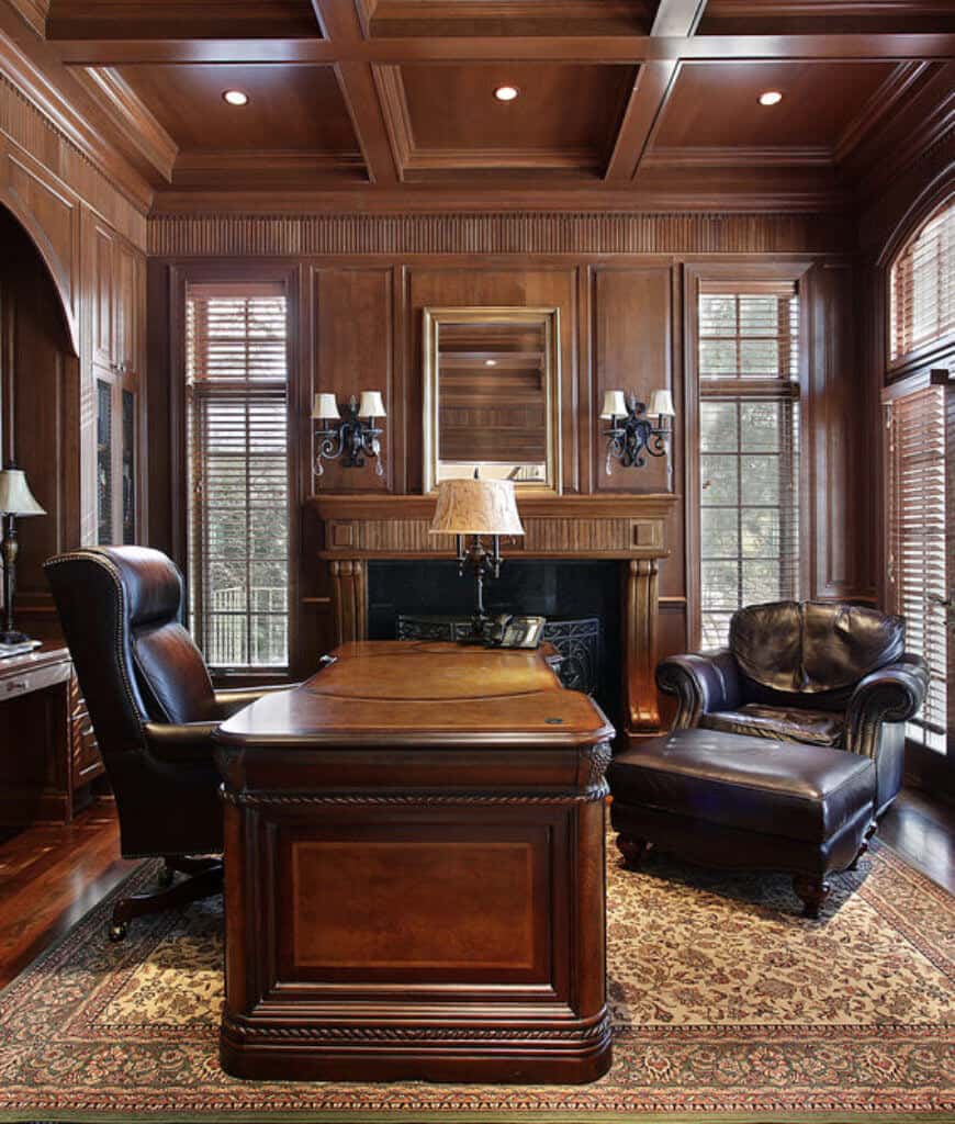 一间全木制的家庭办公室展示了一张木桌，搭配黑色皮椅，复古地毯和壁炉，中间有百叶窗，由锻铁烛台照明。