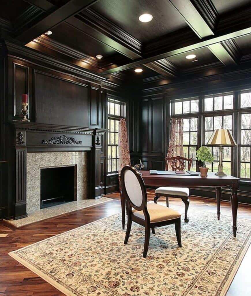 黑色家庭办公室设有一座壁炉以及倾斜布局的木制桌椅。它有格子天花板和木板地板，上面有花卉地毯。