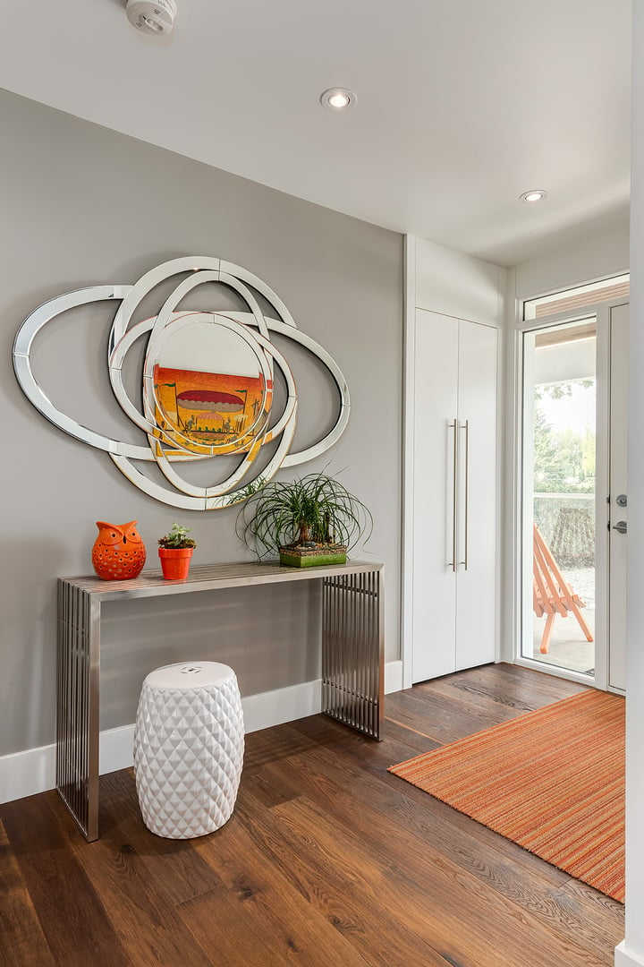 在门厅里，一面镜子悬挂在金属吧台台和白色质感的凳子上。橙色从装饰、条纹地毯和帆布画中突现出来，反射在镜子上。