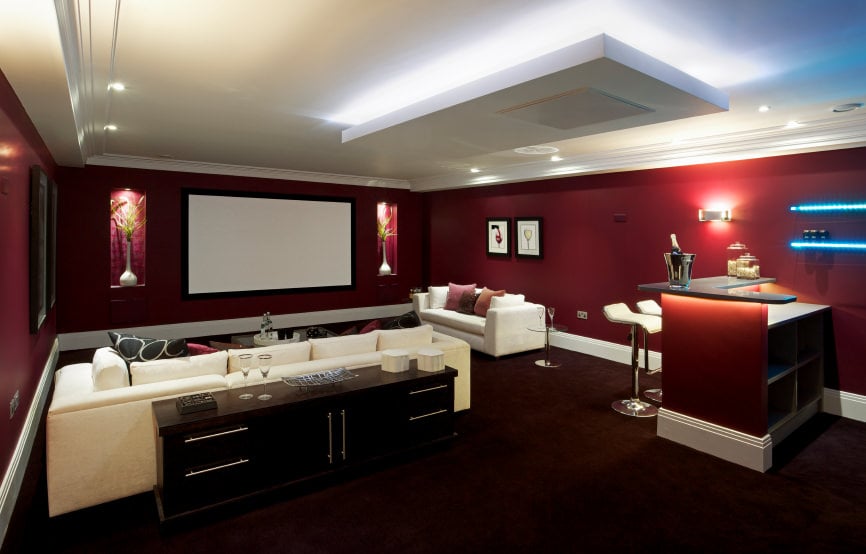 这间现代化的媒体室拥有一个带有组合沙发座椅的剧院空间，旁边的小酒吧设置在红色天鹅绒地毯地板上，与红色优雅的墙壁相匹配。